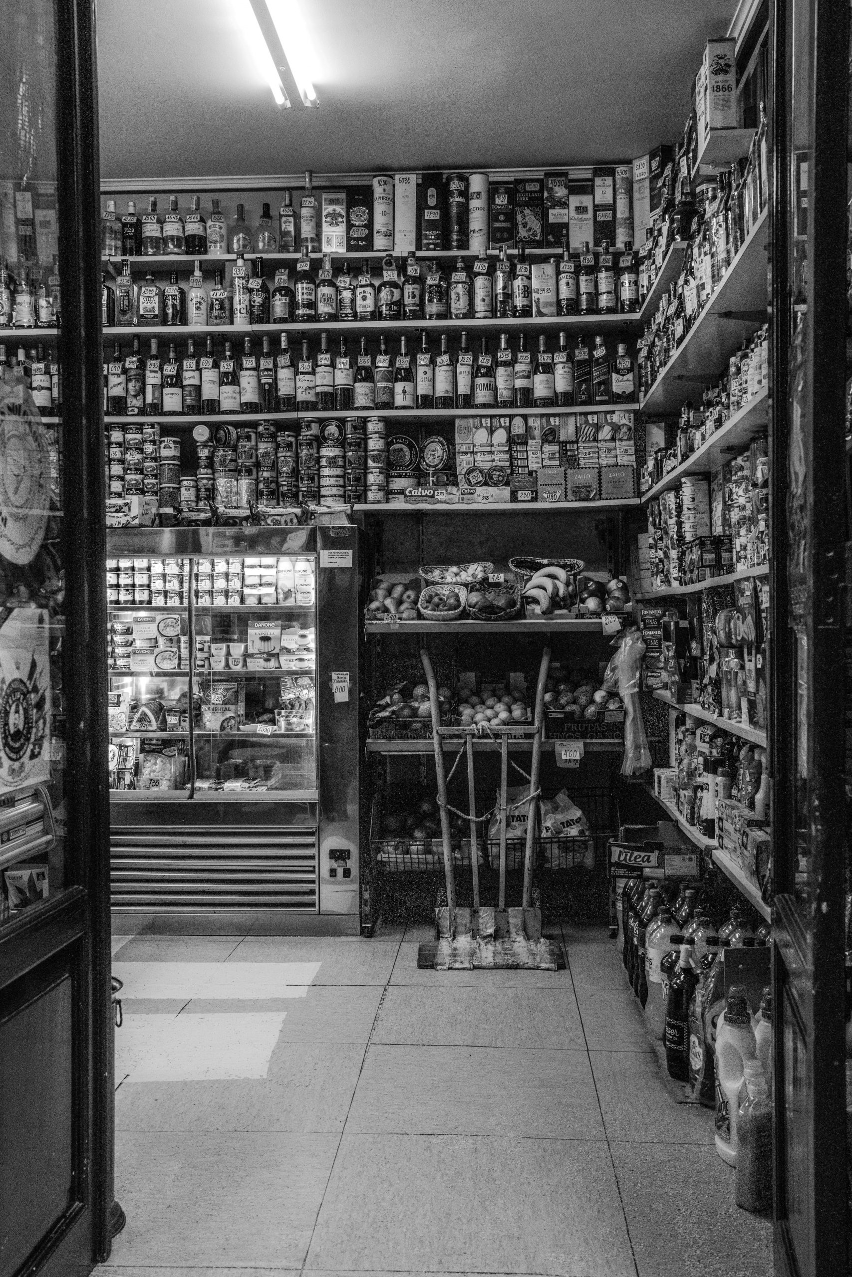 Services Black and white photo of a cluttered grocery store interior filled with various products.